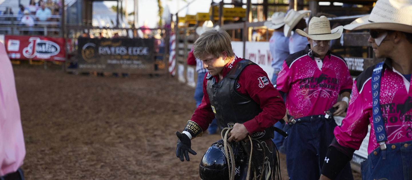 Cowboy wearing a red shirt walking away in the arena holding his helmet.